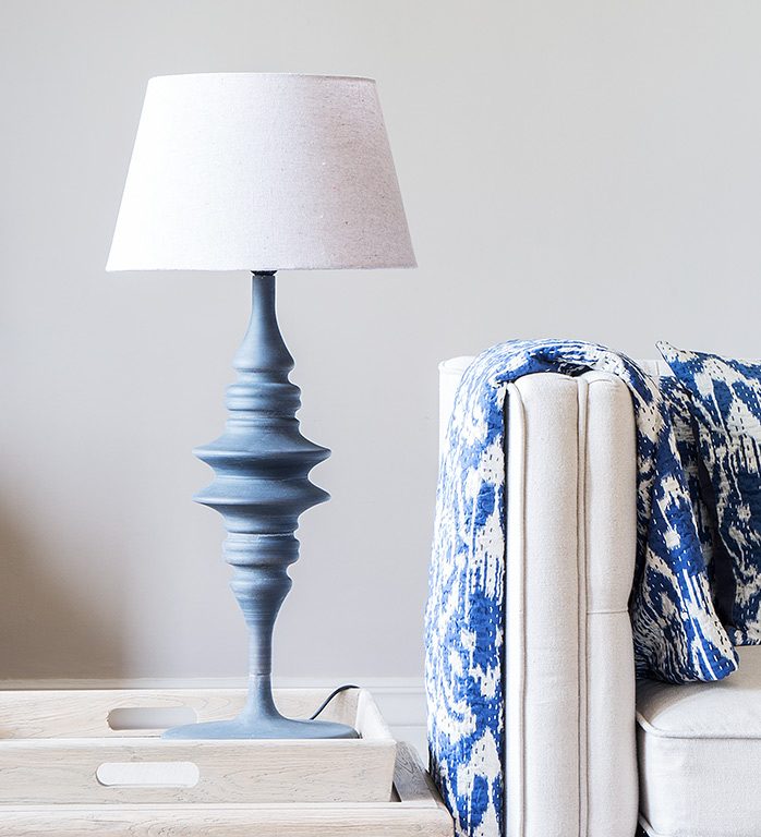 A tall blue/grey metal lamp in with a linen shad stands on a table next to a cream sofa with a blue and white throw
