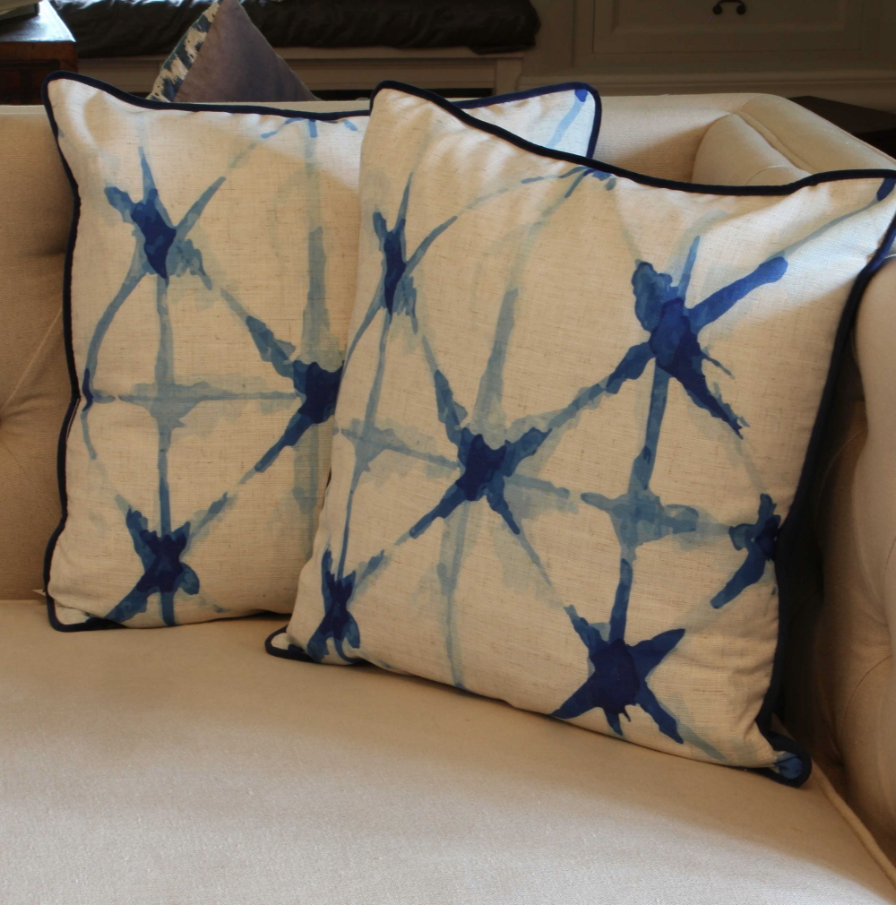 A pair of luxury, square cushions are arranged on a cream sofa. The cushions are cream with printed x detailing in shades of blue and blue trim around the edges.