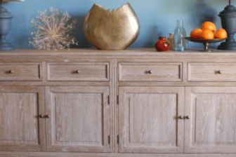 A four door and four drawer cupboard in limed oak is set against a blue wall and has two lamps a gold vase and other decoratives items placed on the top surface
