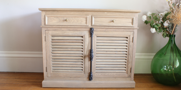New England 600x300 A cupboard with two drawers and two louvred doors sits against a pale wall with a green glass vase to it's right hand side.