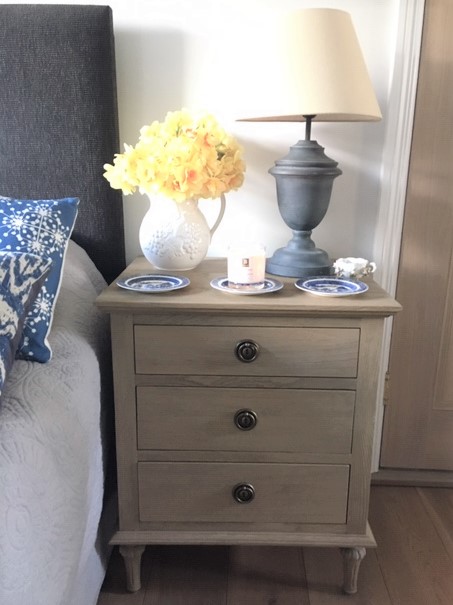 The picture shows the "Melbury" three drawer chest of drawers beside a bed with a grey headboard, white covers and blue and white cushion. On the chest of drawers is a tall grey lamp shaped like an urn, with pale lampshade, a vase of dafodils and some small blue and white china plates, there is a candle on the central plate.