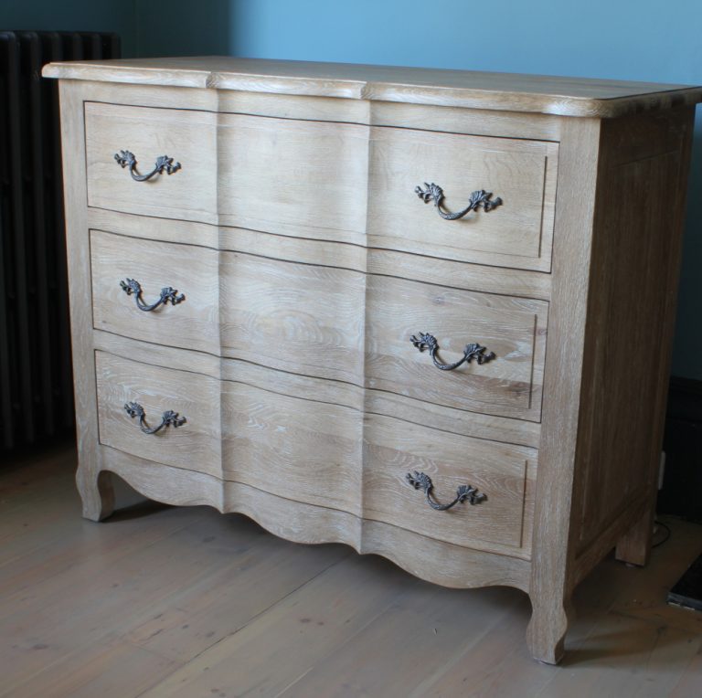 A substantial three drawer chest of drawers with fluted shaping to the front and paired cast iron handles on each drawer stands on a wooden floor in front of a blue wall with strong shadows.
