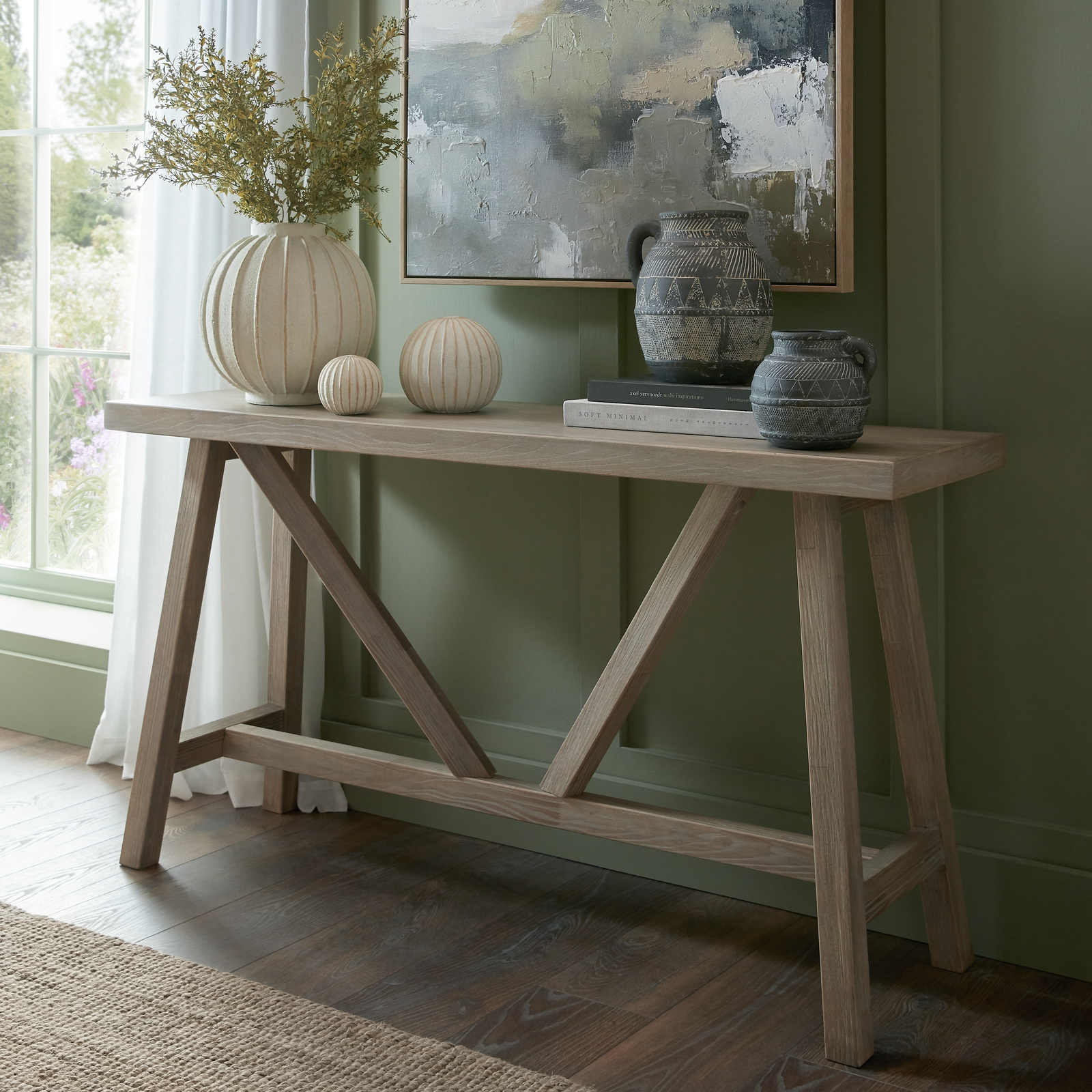 Image shows a wooden console table with a selection of vases and pottery placed on the top. It is next to a large window and there is a television or pice of art on the wall above the table.