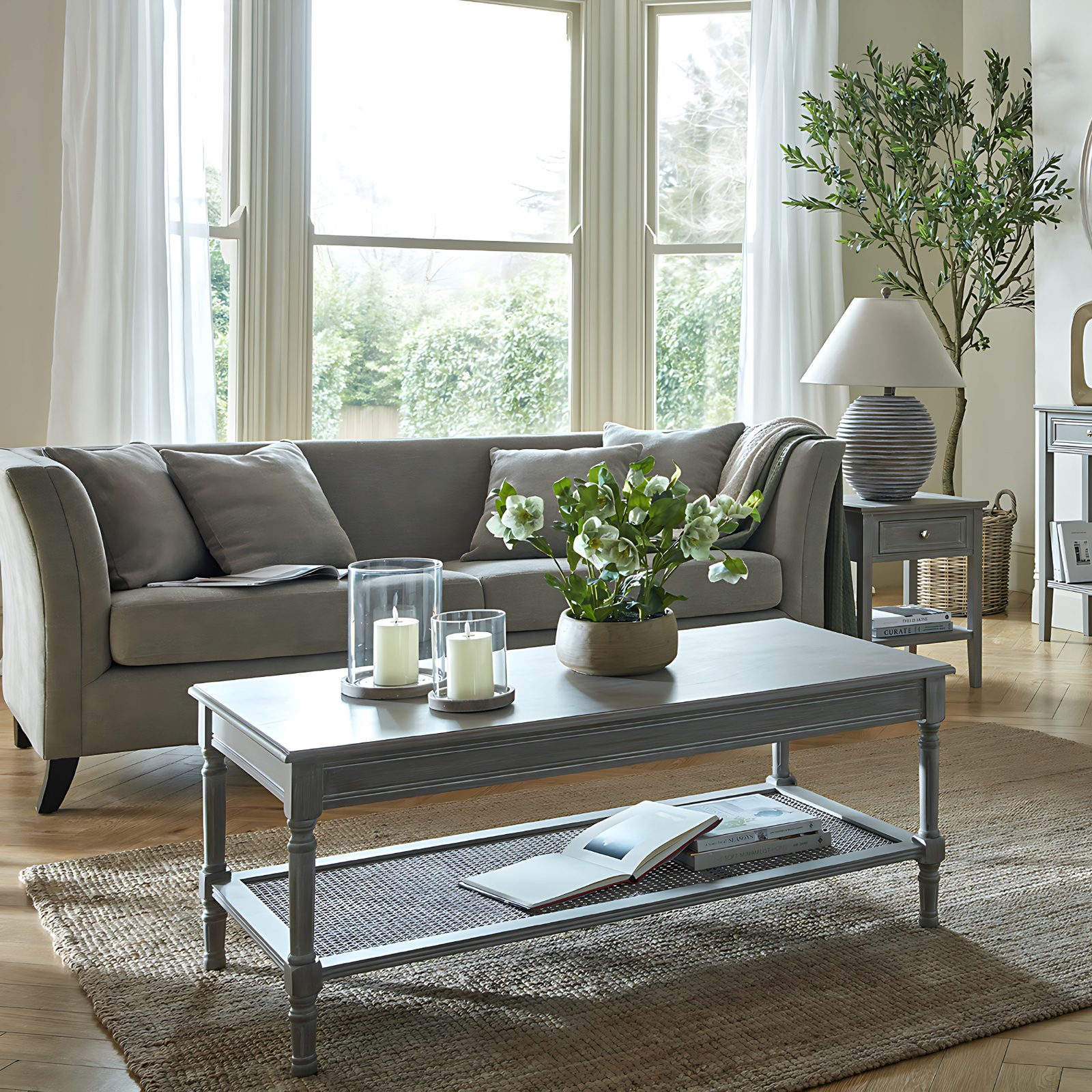 The image shows a sitting room with a coffee table in the foreground with a sofa behind. There is a side table with a lamp on top to the right hand side of the sofa and an indoor tree behind the side table.