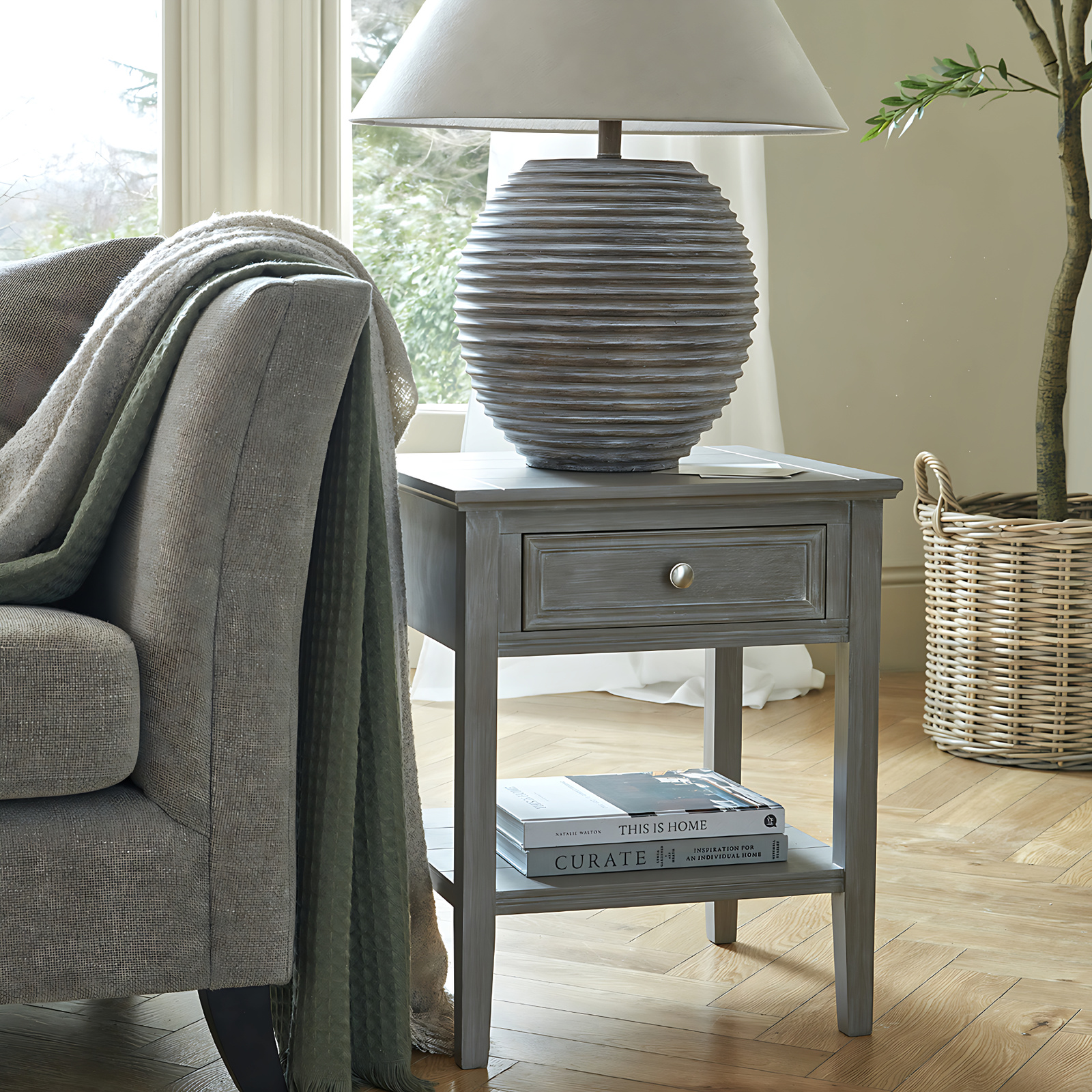 The image shows a wooden side table to the right hand side of the arm of the sofa. there is a throw rug over the arm of the sofa and a round-based table lamp on top of the side table. The side table has drawer at the top with a shelf underneath. there are some magazines/books on the shelf.