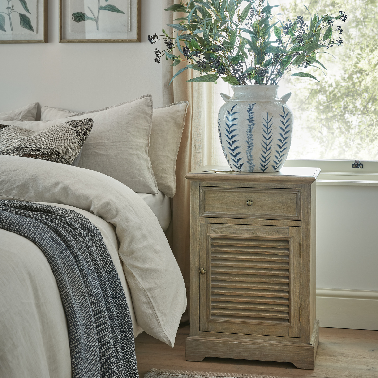 The image shows a brown wood cabinet with one drawer above a single door with the handle on the left and hinges on the right side of the door. It is next to a bed, in front of a window and has a vase with green plant stems in on the top.